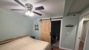 Ein Schlafzimmer mit einem Bett, einem Deckenventilator, einer Holztür und einem durch eine offene Tür einsehbarer Badezimmer.