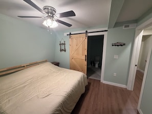 Une chambre à coucher avec un lit, un ventilateur de plafond, une porte en bois et une salle de bain visible à travers une porte ouverte.
