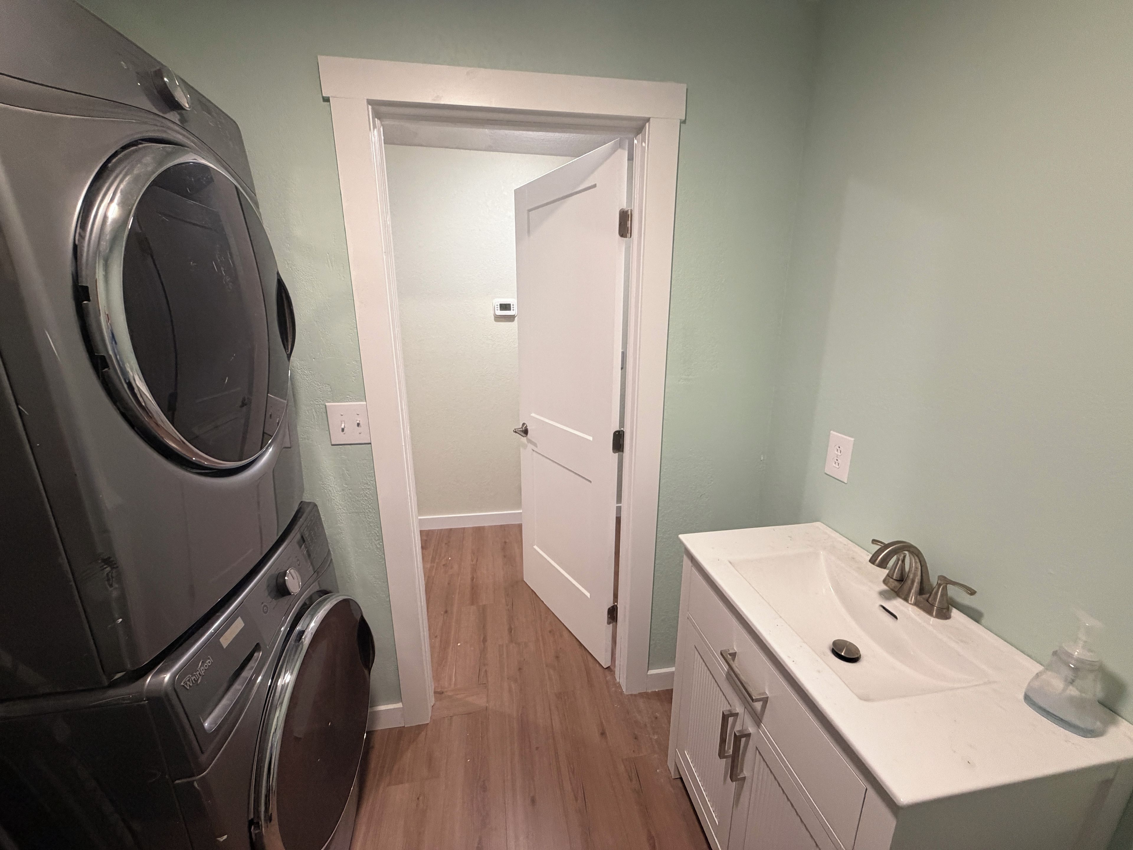 A laundry area with a front-loading washer and dryer, a white vanity with a sink, and a door leading to a hallway.