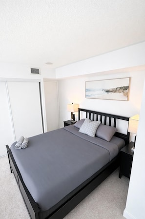 A neatly made bed with a grey mattress and pillows, a black headboard, two bedside tables with lamps, a white wall with a framed picture, and a white door. - CityPlace Condo Suites near the Lake (Toronto)