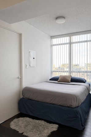 A bedroom with a bed, a white door, a window with blinds, and a view of buildings outside. - CityPlace Condo Suites near the Lake (Toronto)