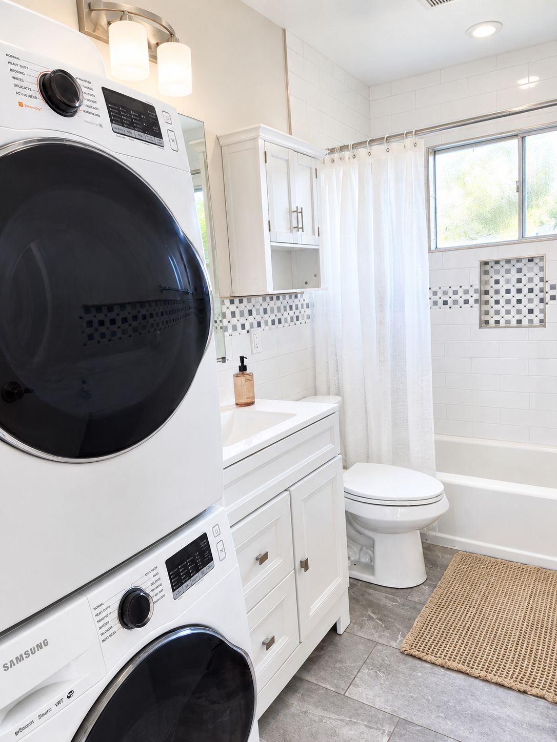 Un área de lavandería con lavadoras y secadoras apiladas, un baño con inodoro, bañera y ventana.