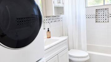 Un área de lavandería con lavadoras y secadoras apiladas, un baño con inodoro, bañera y ventana.