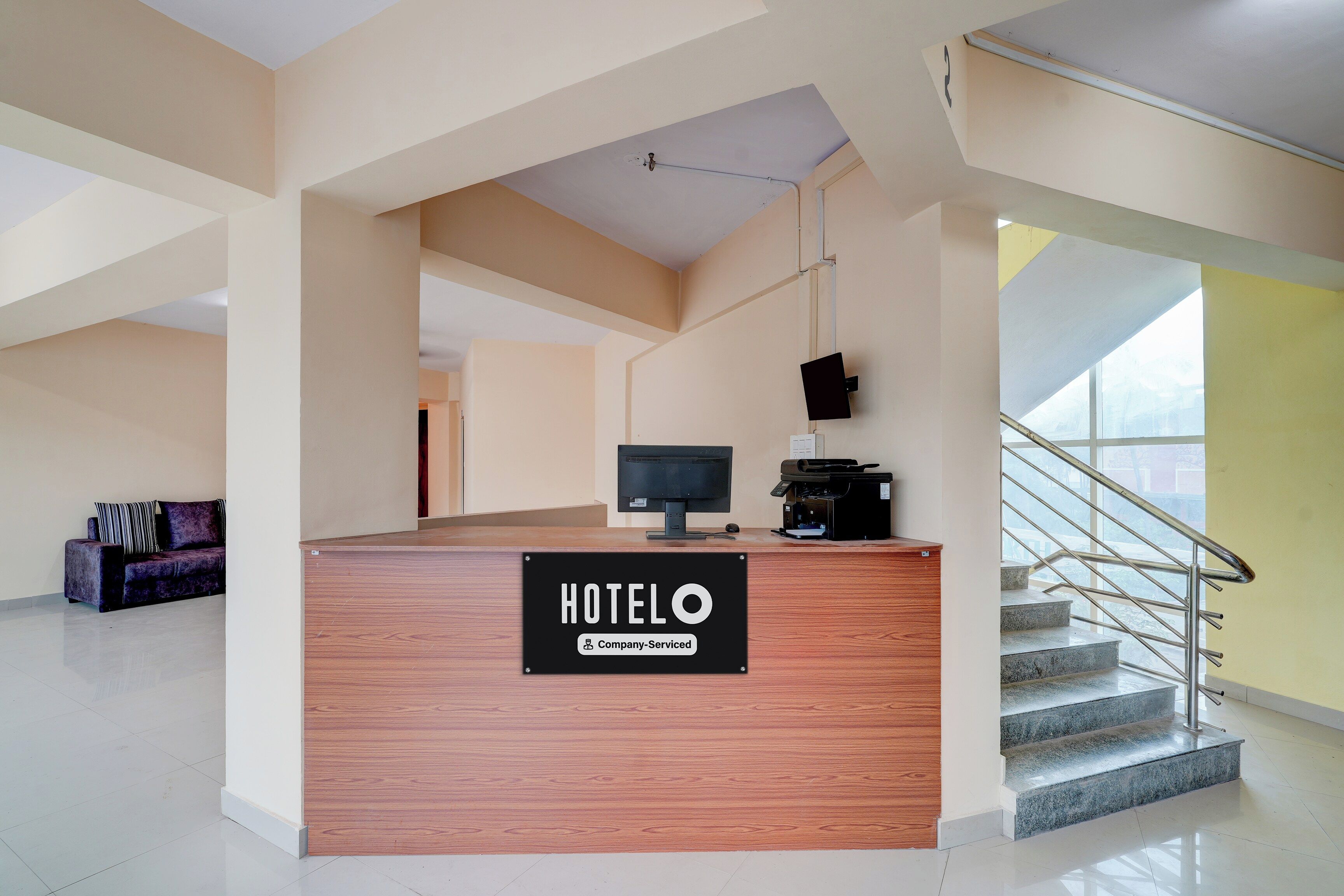 A hotel reception desk with a sign indicating it is company-serviced. There is a computer monitor, a printer, and a sofa in the background.