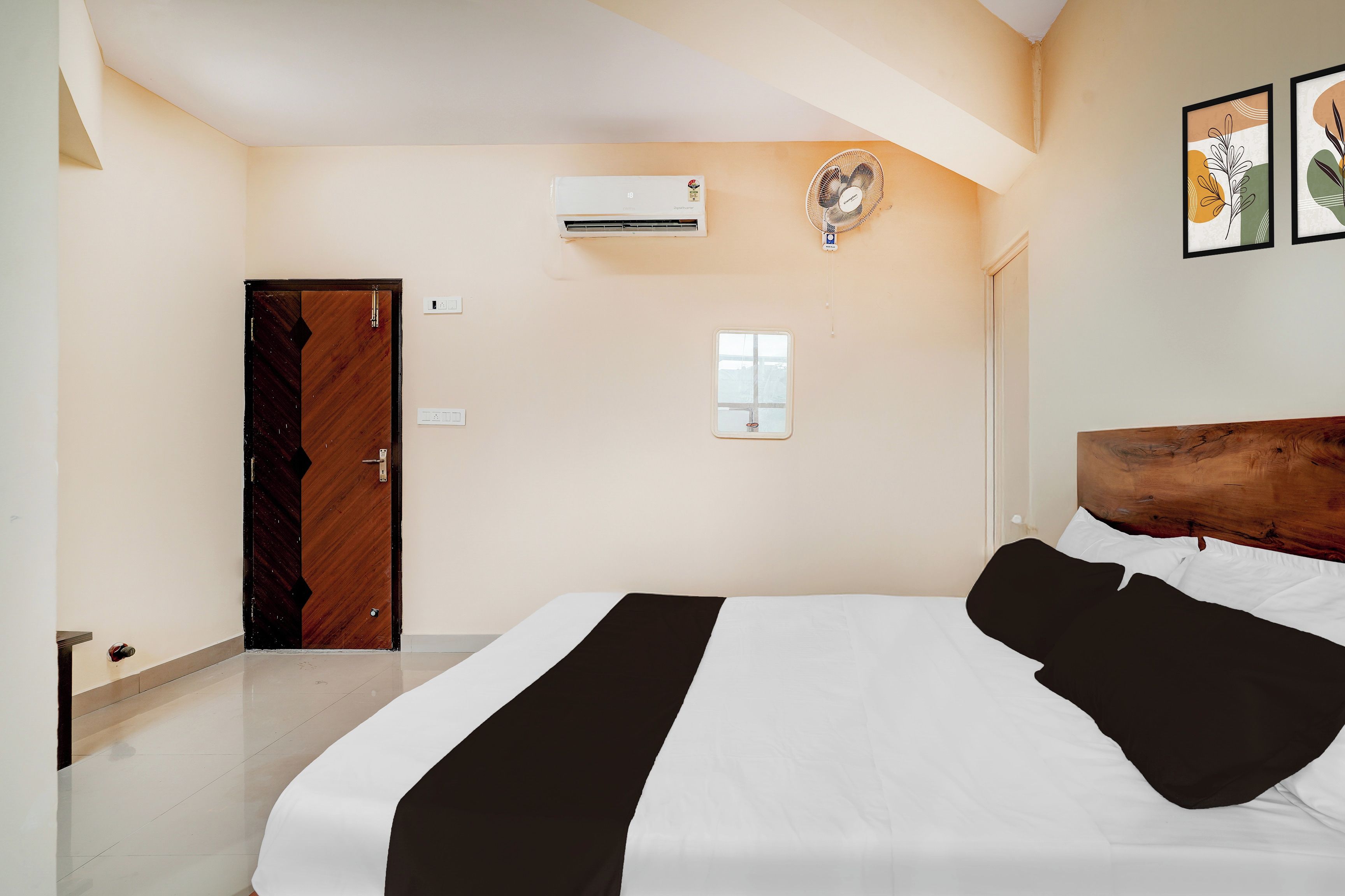 A hotel room with a bed, a wooden door, an air conditioning unit, a small window, and a wall-mounted fan.