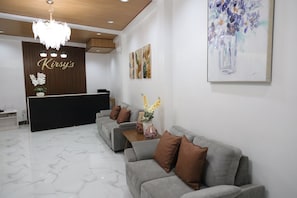 Una zona de recepción con mostrador, una araña de cristal, un sofá gris y un cuadro en la pared.