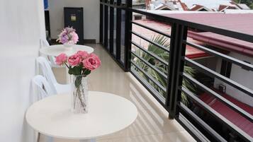 A balcony with a white table and chairs, a vase with pink flowers, and a view of buildings and greenery.