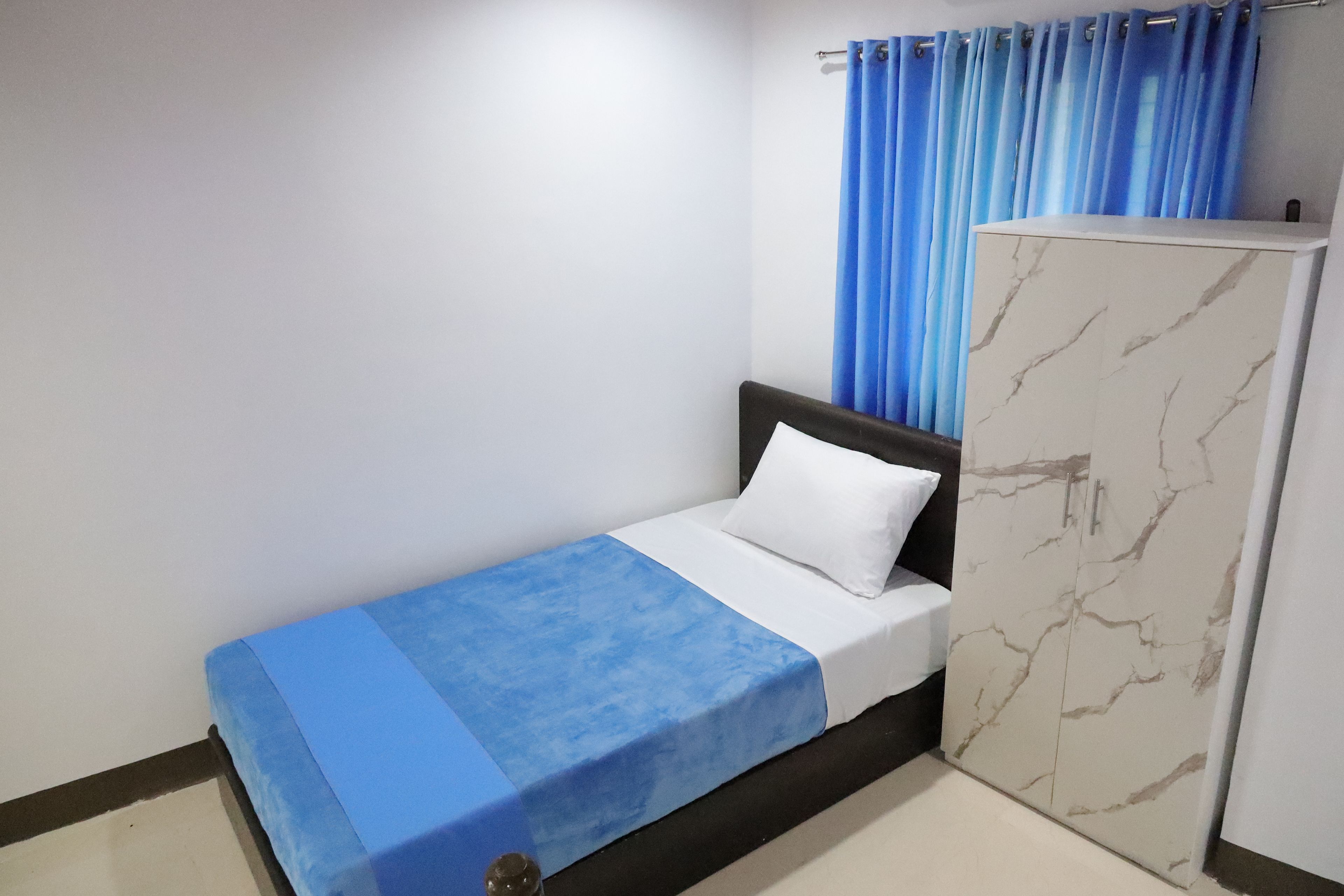 Una cama individual con un almohadón blanco y una manta azul, un armario con estampado de mármol y cortinas azules en una ventana.