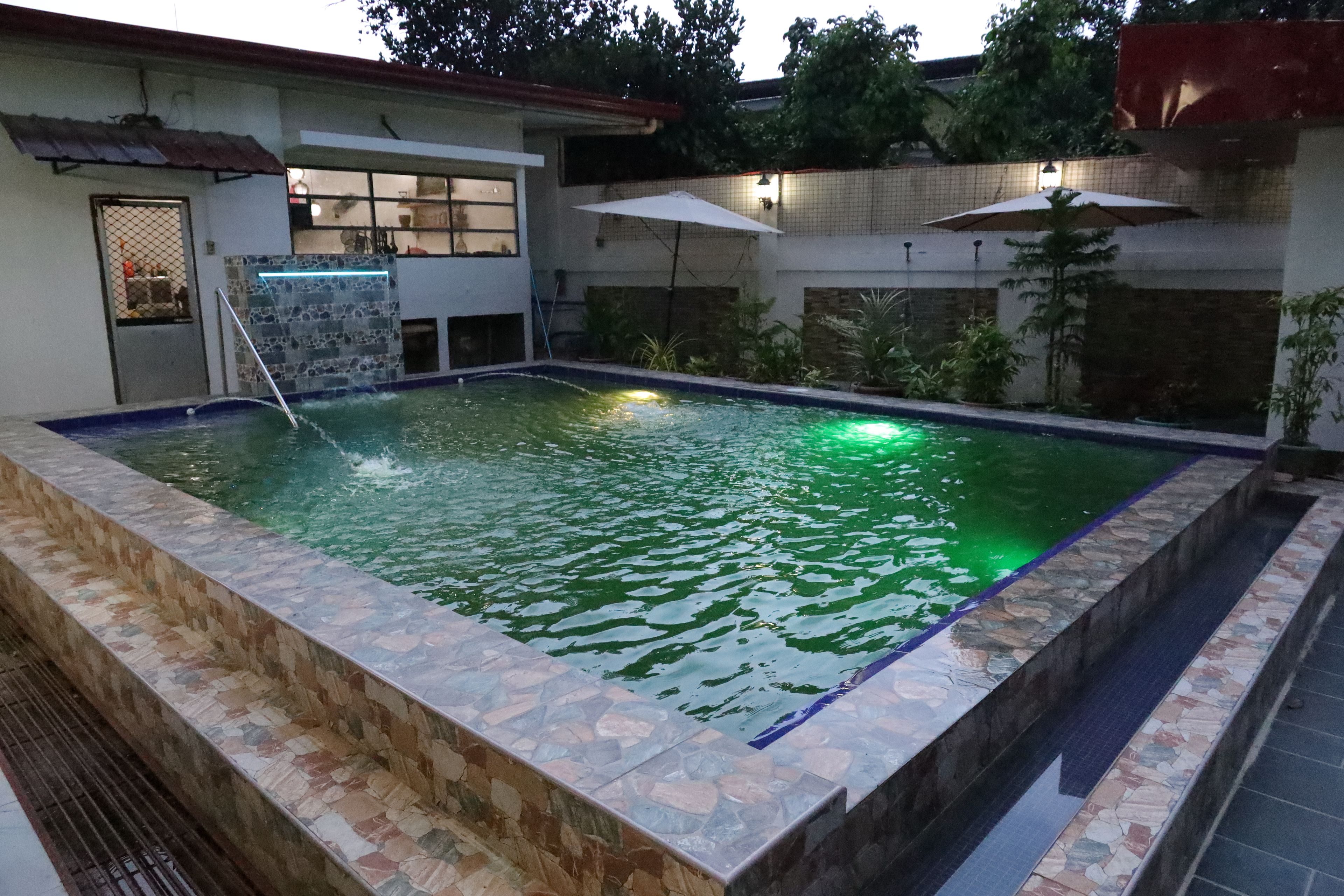 A swimming pool with a stone border, surrounded by a building with a tiled roof and a door with a mesh screen.