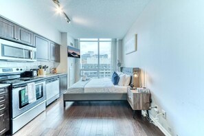 A compact bedroom with a bed, bedside table, and a window with a city view. - Fort York Condos near the Waterfront (Toronto)