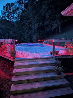 A wooden deck leading to a hot tub area with a pool, surrounded by trees and illuminated by red lighting.