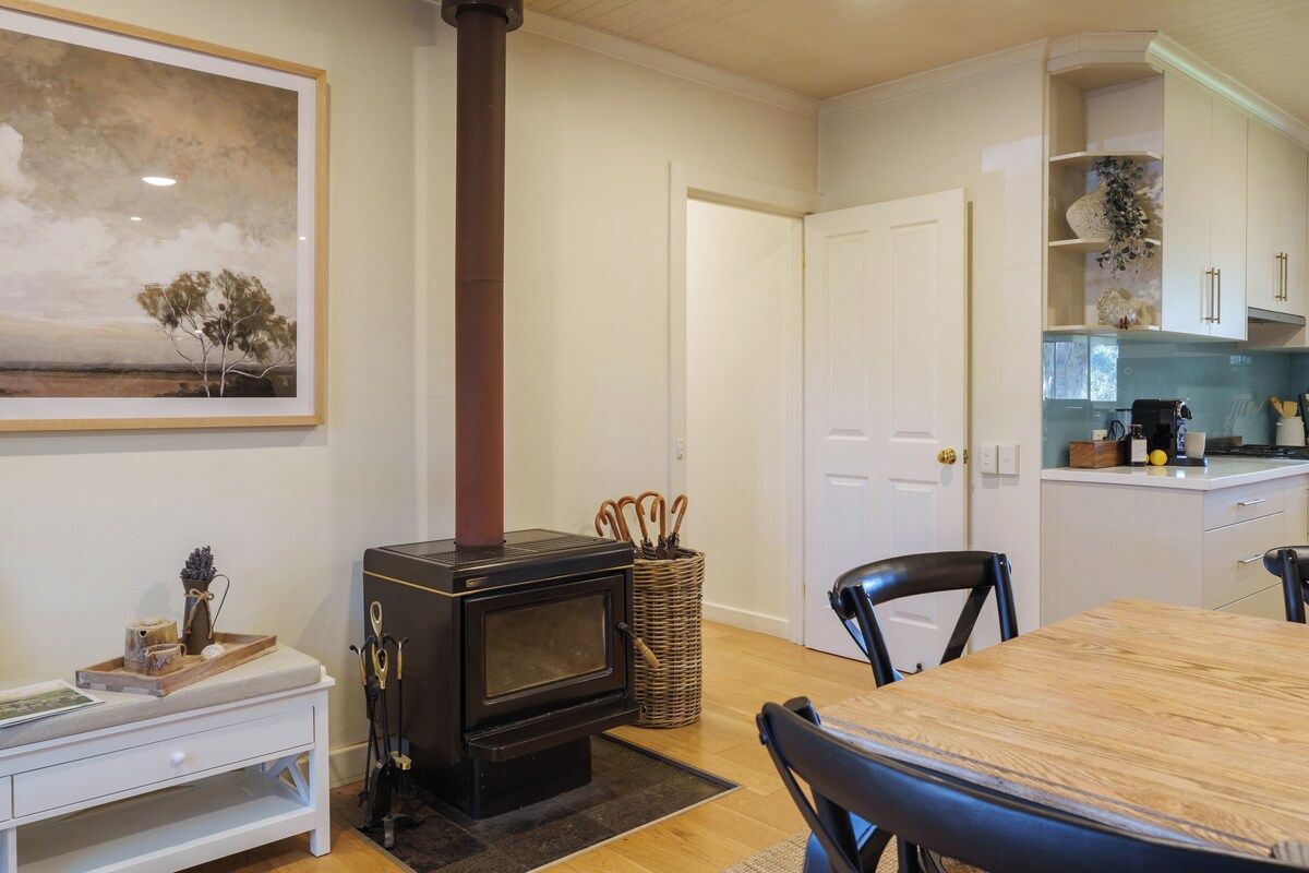 Une cuisine chaleureuse avec un poêle à bois, une table à manger avec des chaises et un meuble blanc agrémenté d’objets décoratifs.