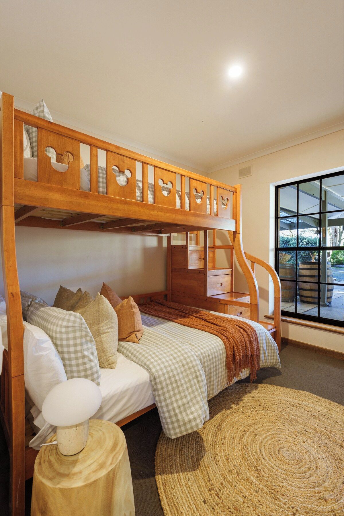 A bunk bed with a wooden headboard featuring cut-out Mickey Mouse ears, a bedside table with a lamp, and a view of the outdoors through a large window.