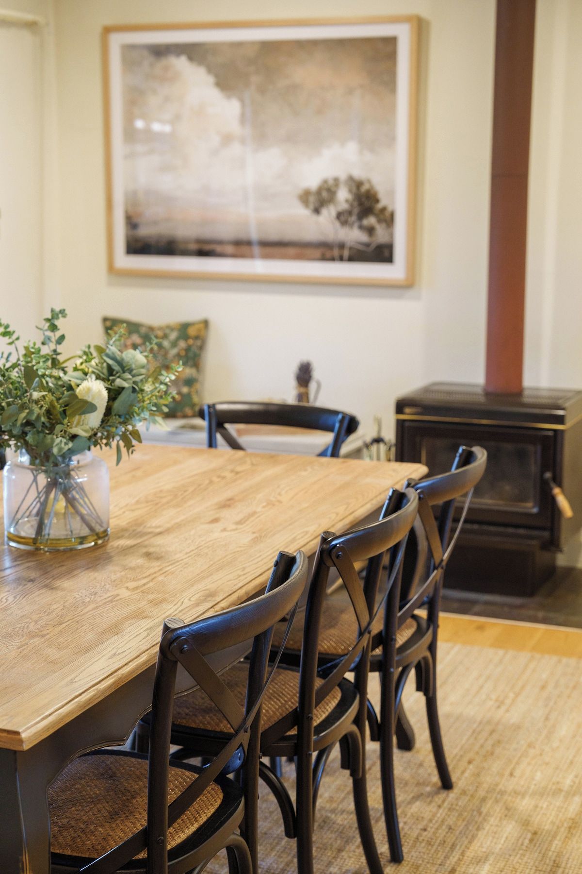 A dining area with a wooden table, black chairs, a vase with flowers, and a painting on the wall.