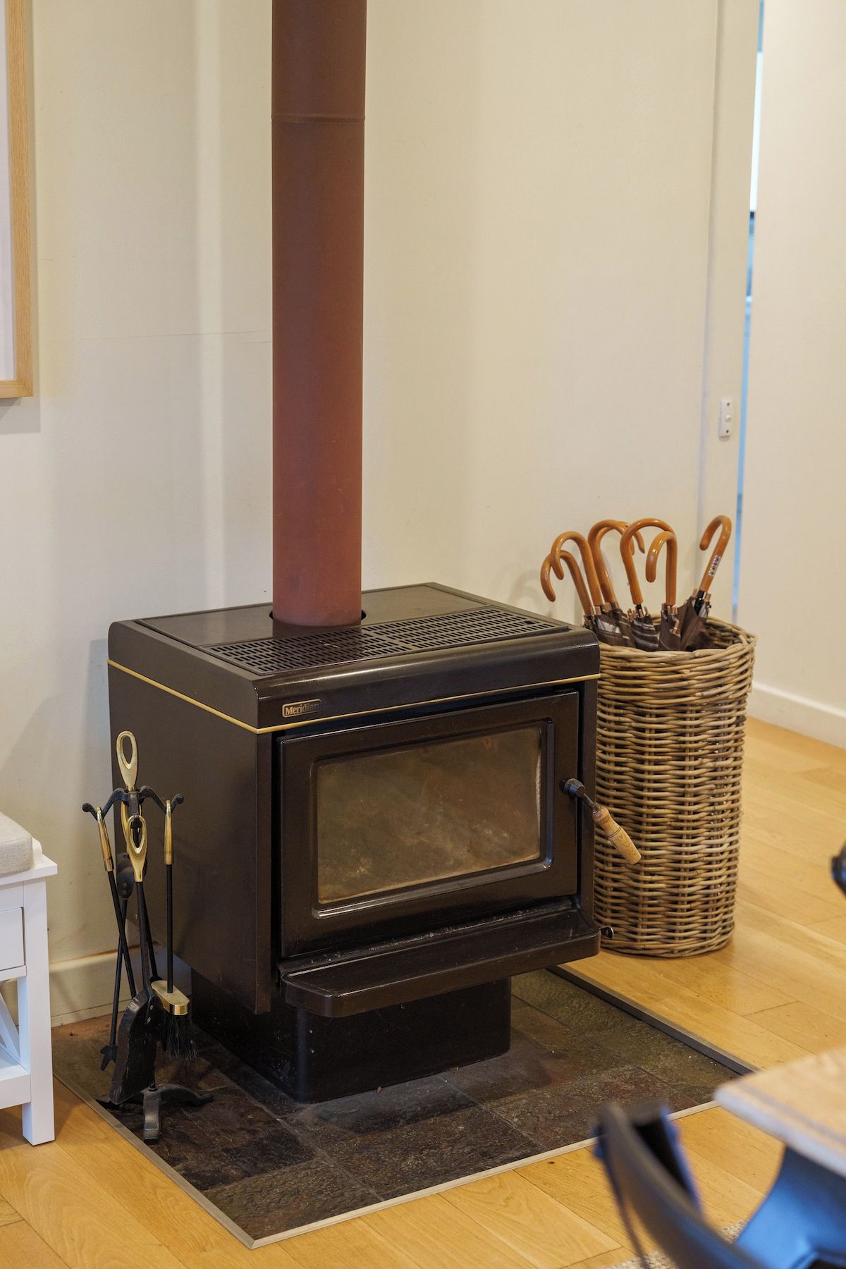 A wood-burning stove with a chimney, a wicker basket with utensils, and a small white stool.