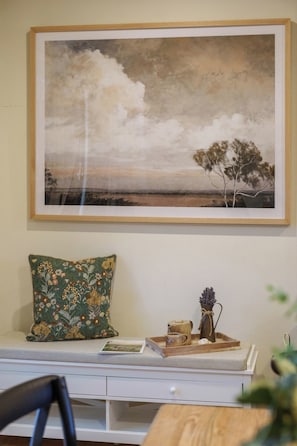 A framed landscape painting above a white console table with a floral pillow, a tray with a mug, and a vase with flowers.