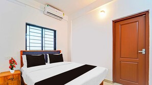 Une chambre d’hôtel avec un lit, une fenêtre, un appareil de climatisation, une porte en bois et une table de chevet ornée d’un vase de fleurs.