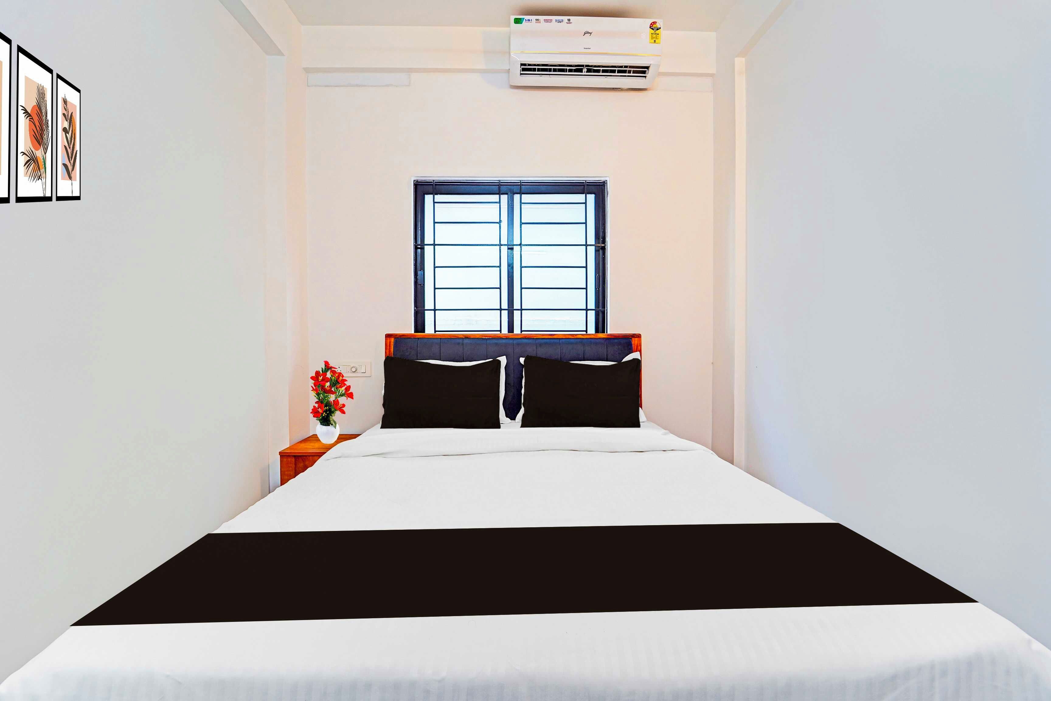 A bedroom with a bed, a window, an air conditioning unit, and a nightstand with a vase of flowers.