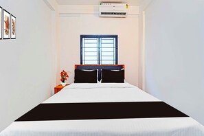 Un dormitorio con cama, ventana, equipo de aire acondicionado y mesita de noche con un florero.