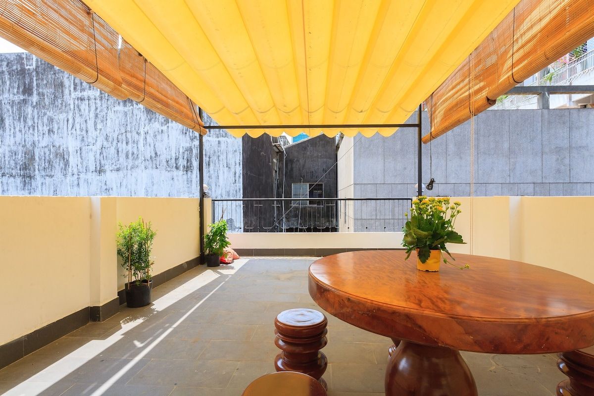 An outdoor patio with a round wooden table, potted plants, and a yellow canopy.