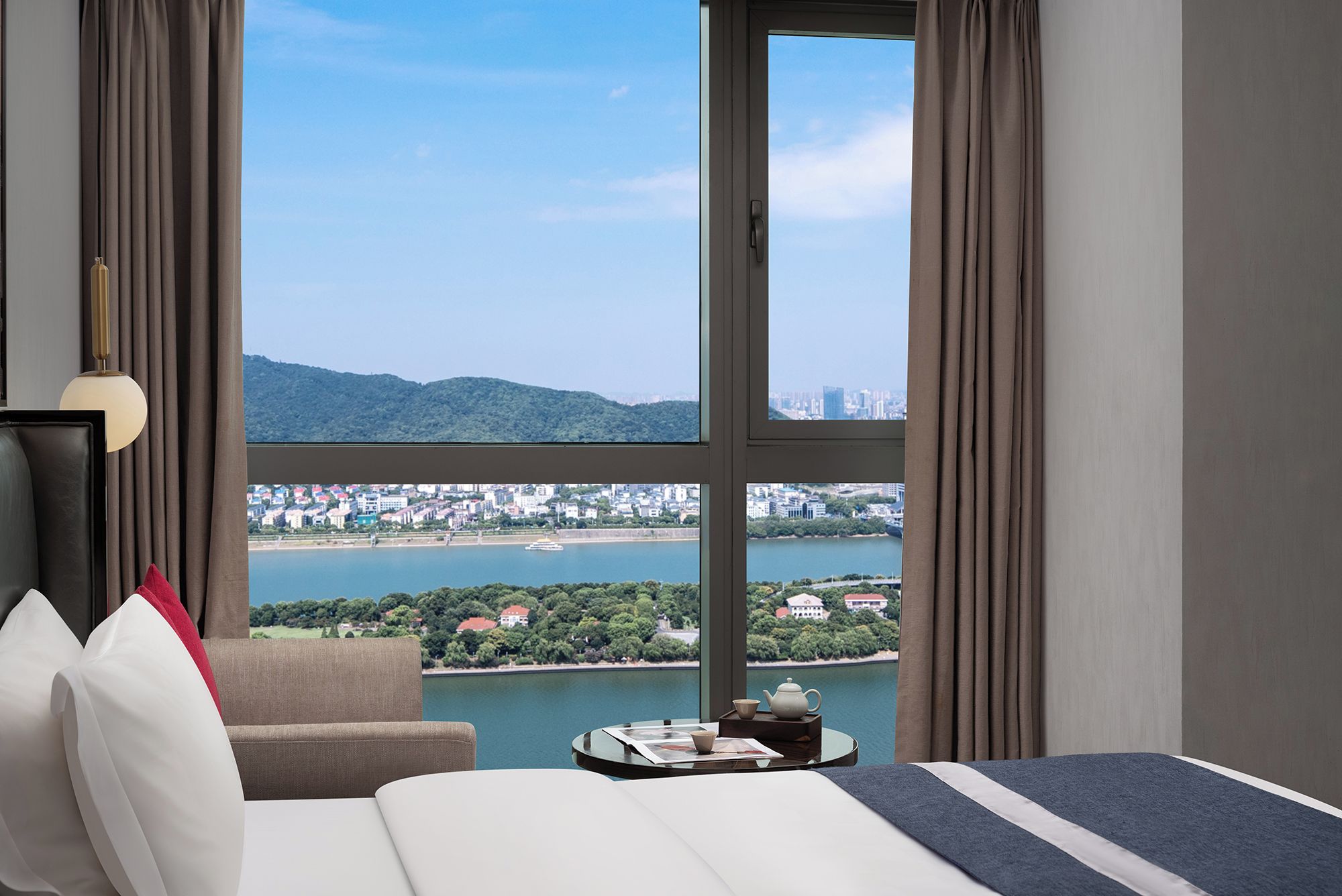Habitación de hotel con un ventanal grande, una cama, una mesita de noche con tetera y vistas a la ciudad y al agua.
