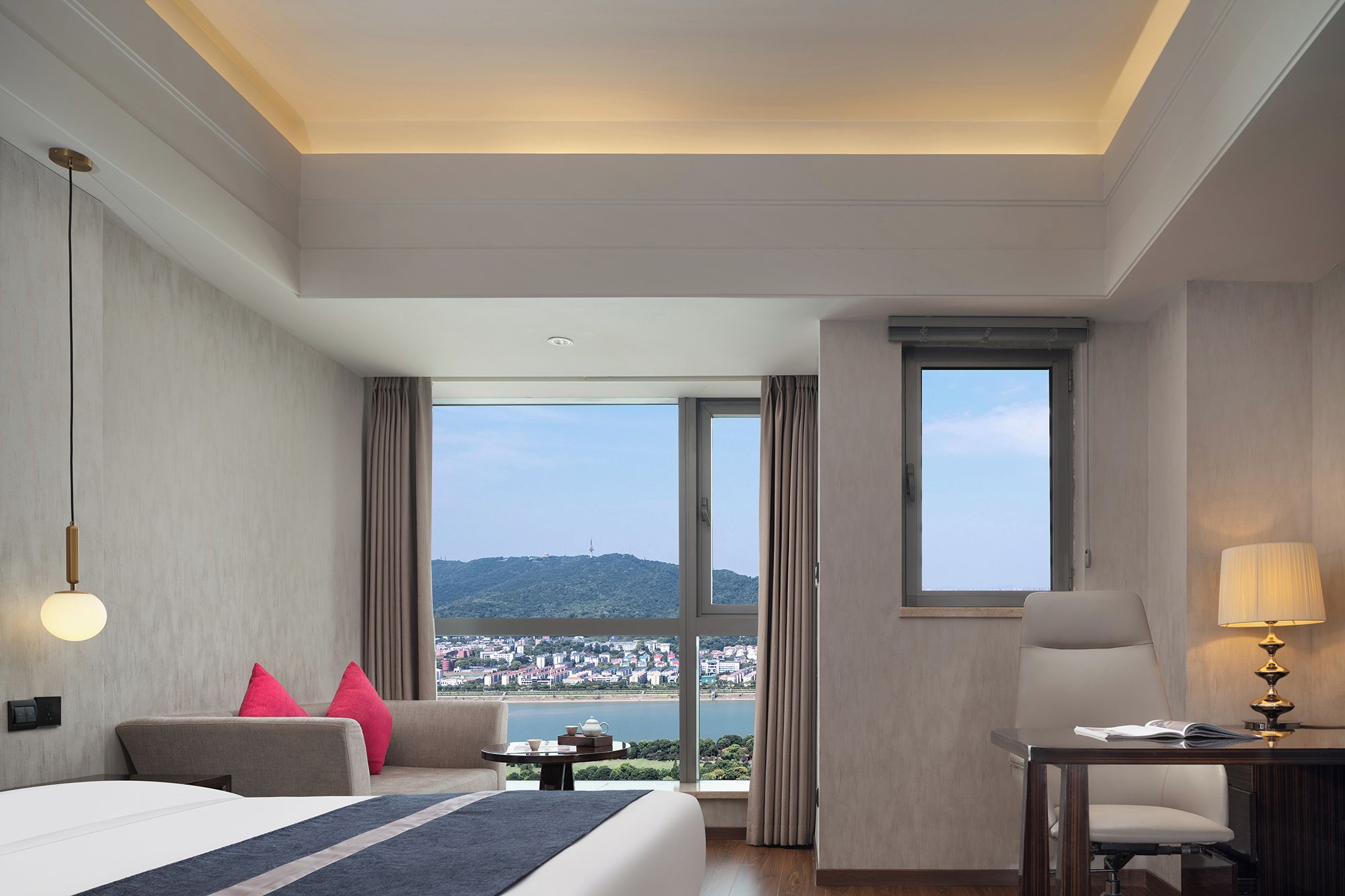 A hotel room with a bed, a desk, a chair, a lamp, and a view of a cityscape through large windows.