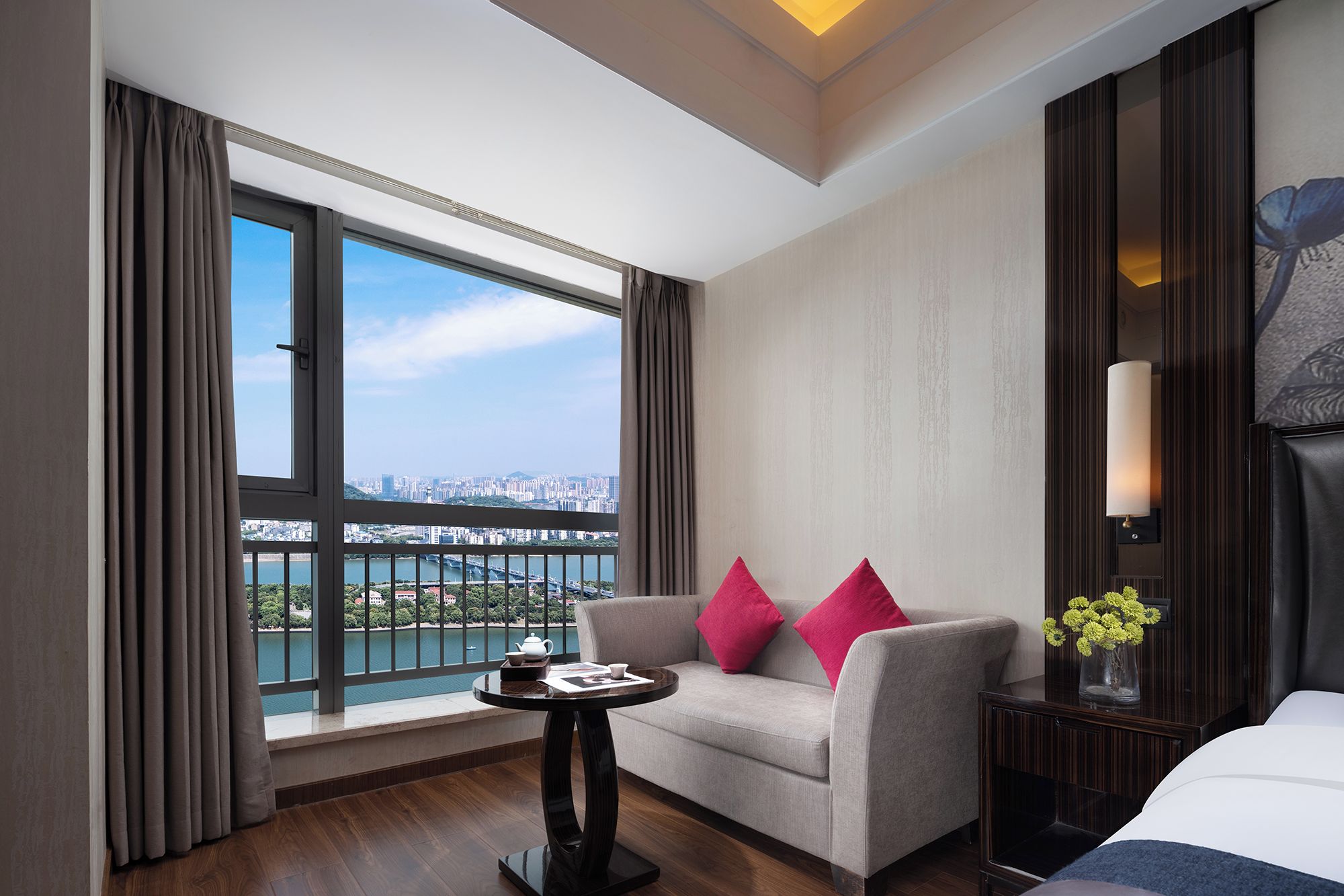 A hotel room with a sofa, a small table, a lamp, and a view of a cityscape through a large window.