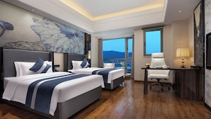 Ein Hotelzimmer mit zwei Betten, einem Schreibtisch, einem Stuhl und einem großen Fenster mit Blick auf die Stadt.