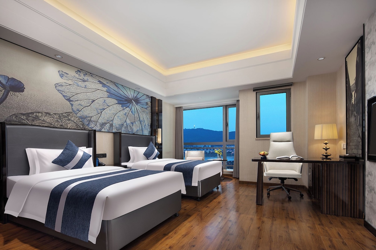 Habitación de hotel con dos camas, un escritorio, una silla y un amplio ventanal con vistas a la ciudad.
