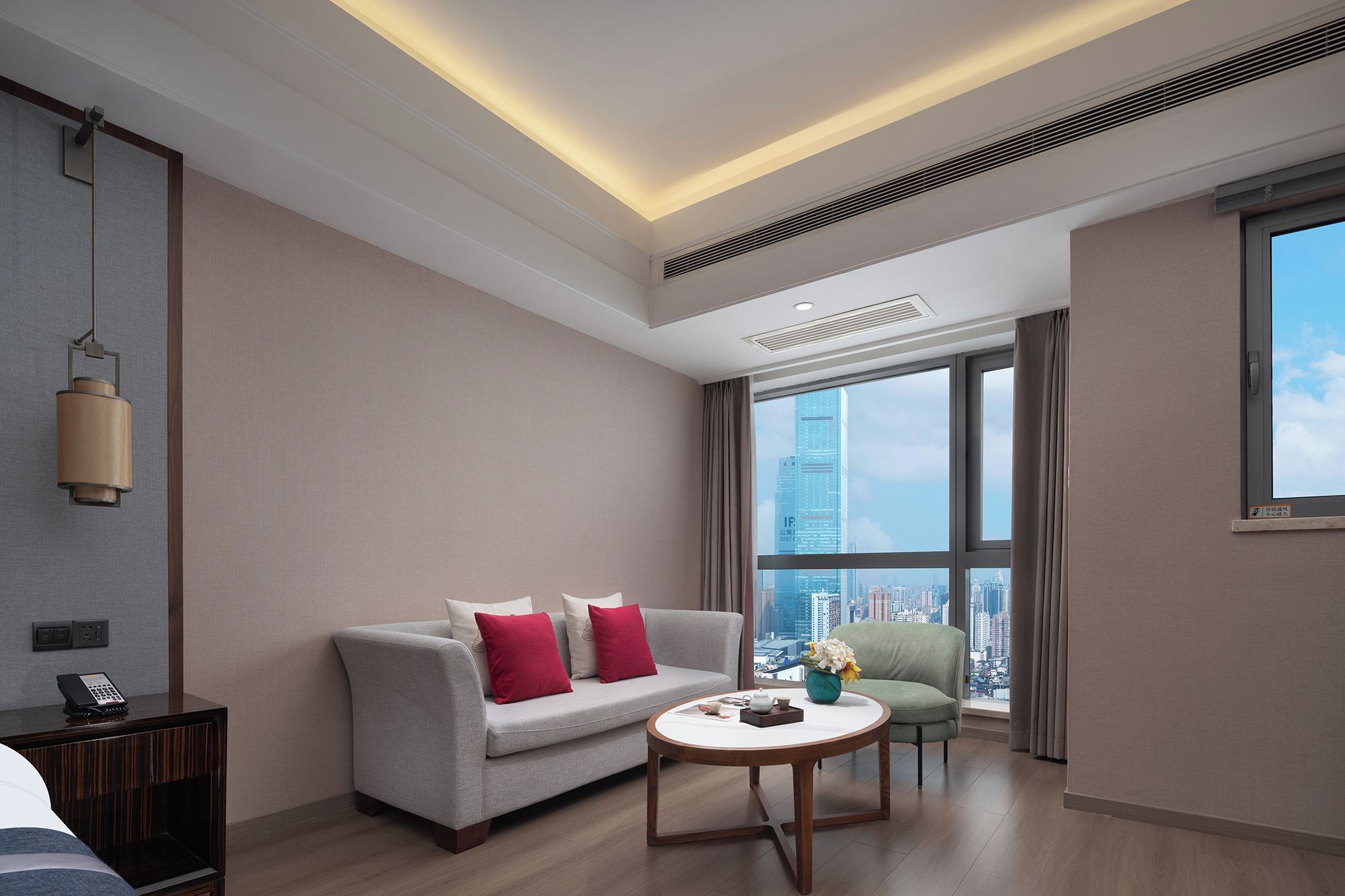 A modern hotel room with a sofa, a round coffee table, and a city view through large windows.