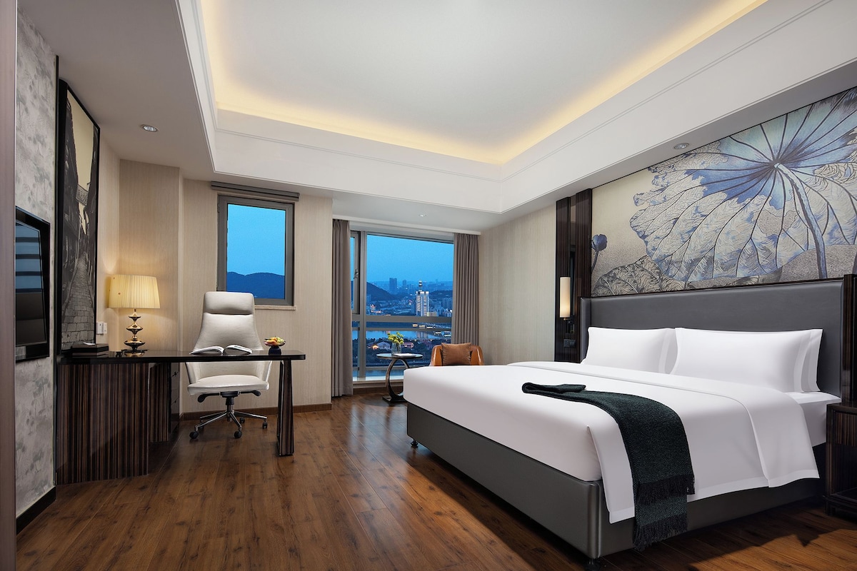 Habitación de hotel con cama, escritorio, silla, lámpara y un amplio ventanal con vista a la ciudad.
