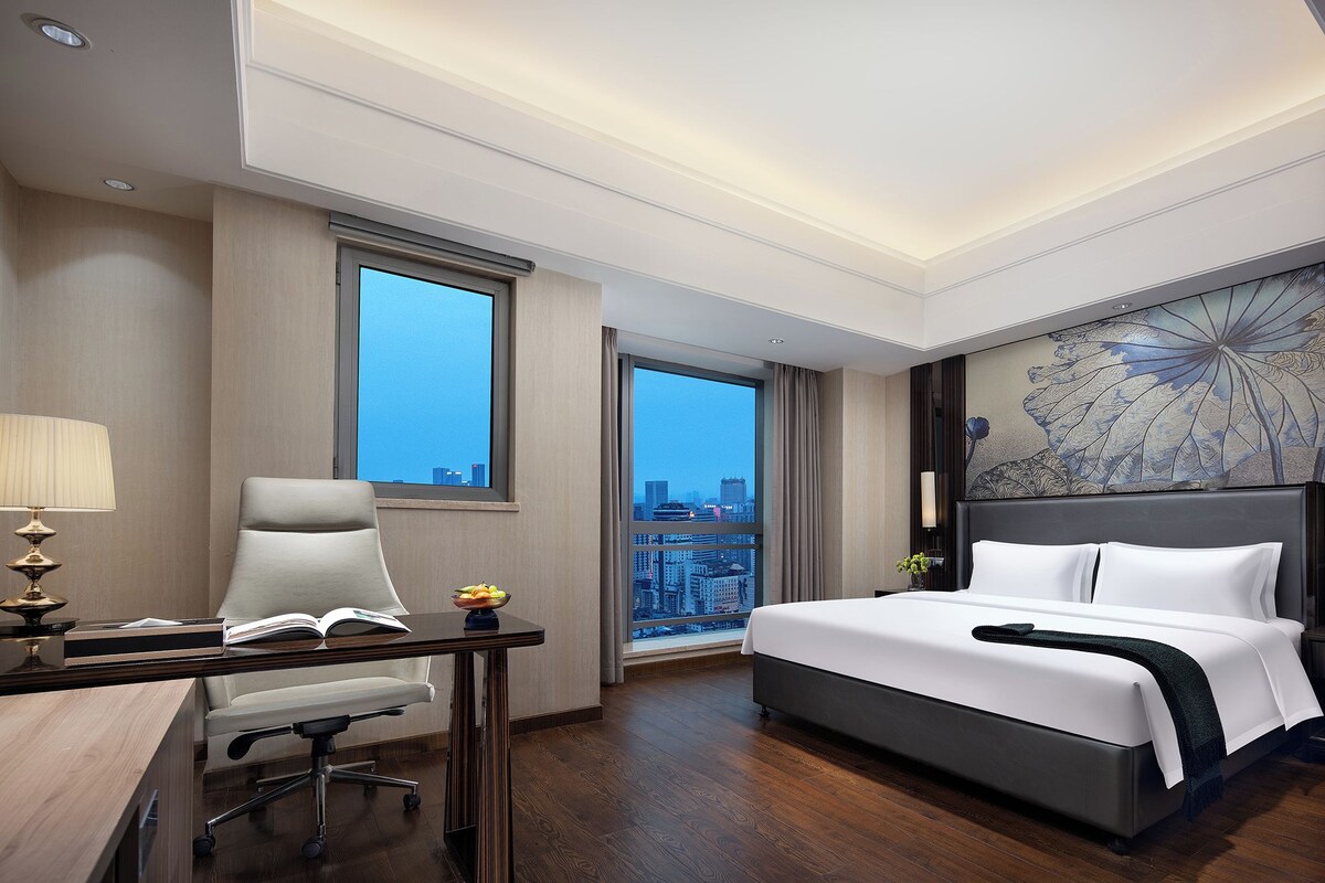 Habitación de hotel con cama, escritorio, silla y ventanas amplias con vistas a la ciudad.