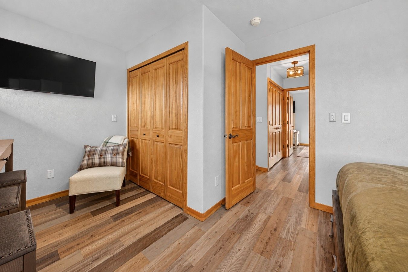 A bedroom with a wooden wardrobe, a bed, a chair, and a television on the wall.