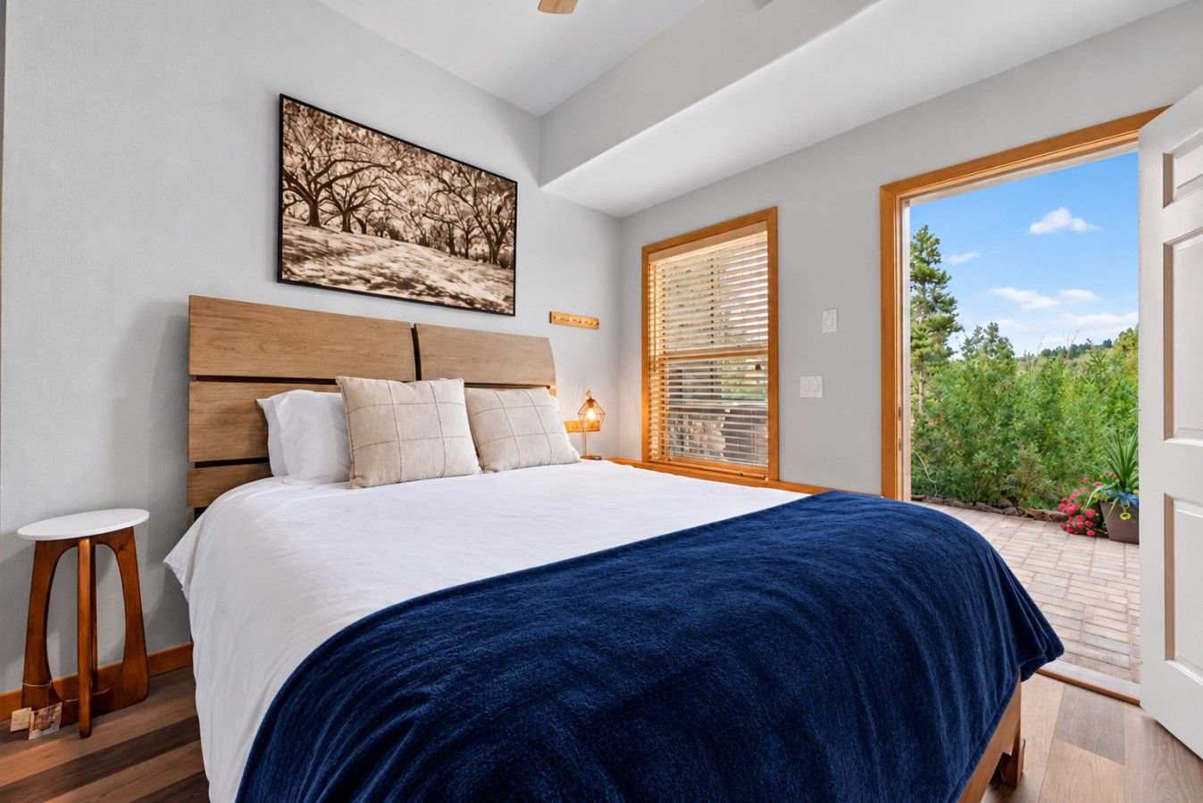 A bedroom with a large bed, a nightstand, a bedside lamp, a window with blinds, and a view of greenery outside.