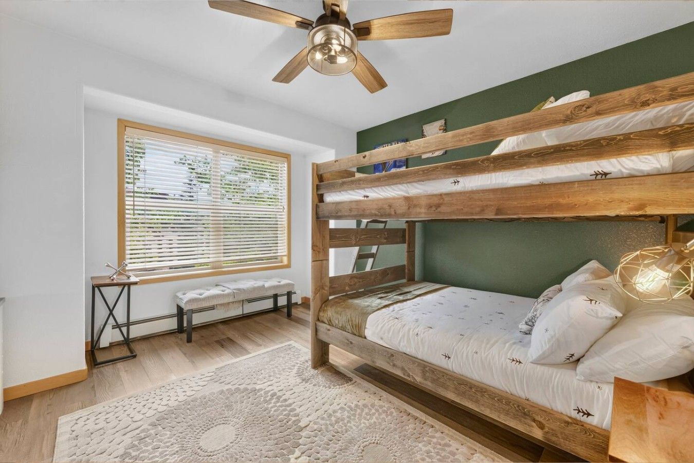 A bunk bed room with a ceiling fan, a window with blinds, a bench, and a patterned rug.