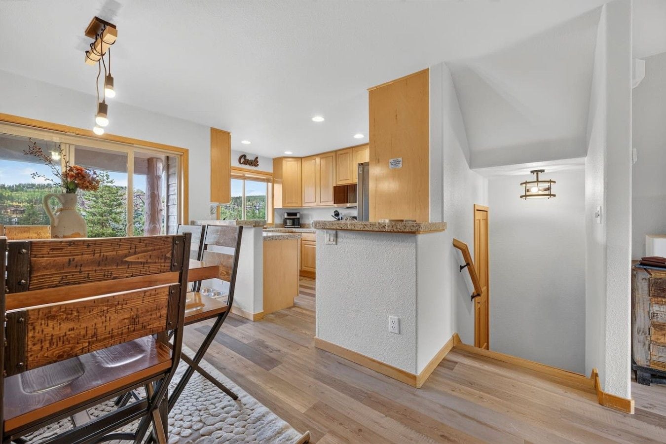 A modern kitchen with wooden cabinets, a dining area with a wooden table and chairs, and a view of the outdoors through a window.