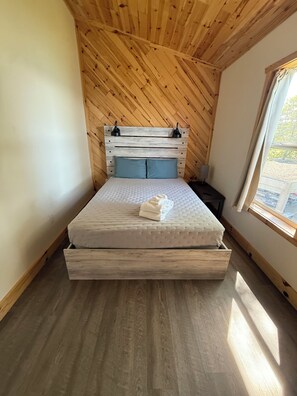 Ein Schlafzimmer mit Holzbalkendecke, einem Bett mit Matratze und Kissen, einem Nachttisch und einem Fenster mit Vorhängen.