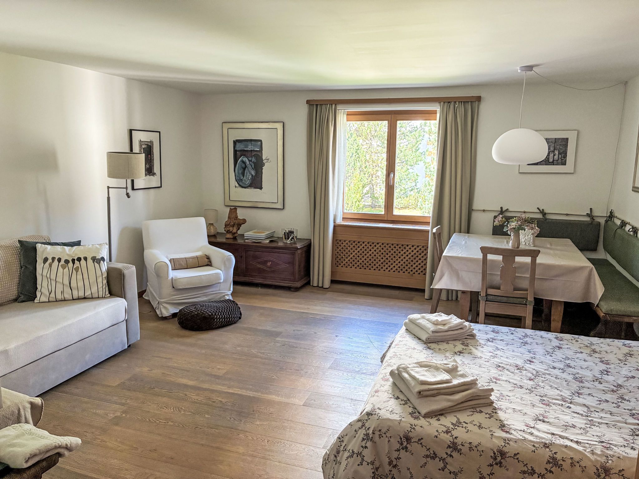 Um quarto com cama, poltrona, mesa e janela com cortinas.