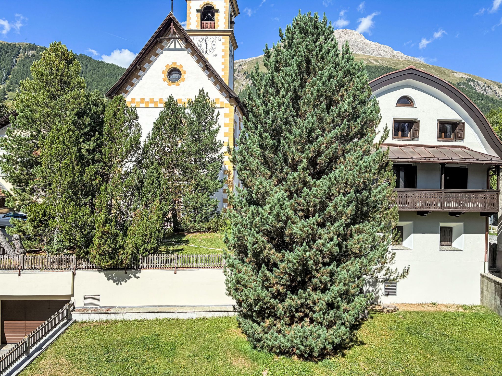 Une église dotée d’un clocher, entourée de pins et avec une montagne en arrière-plan.