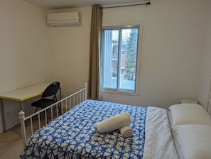 Une chambre à coucher comprenant un lit, un bureau, une chaise, une fenêtre donnant sur des immeubles et un climatiseur.