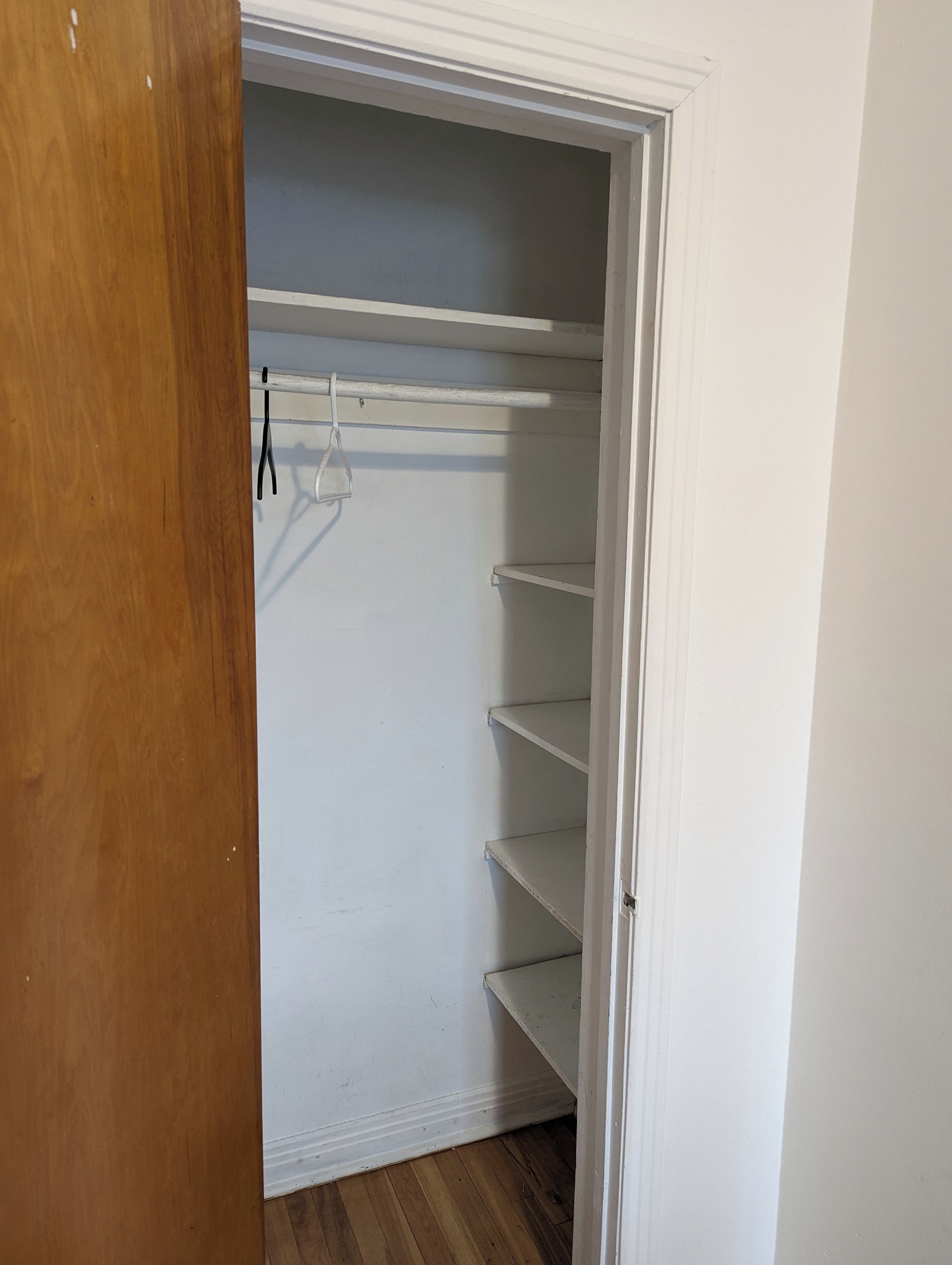 A closet with white walls and wooden flooring, featuring a hanging rack and multiple shelves.