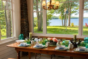 A dining table set with plates, cups, and a vase of flowers, overlooking a lake with trees and chairs.