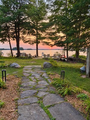 A stone pathway leading to a lakeside seating area with chairs and a grill.
