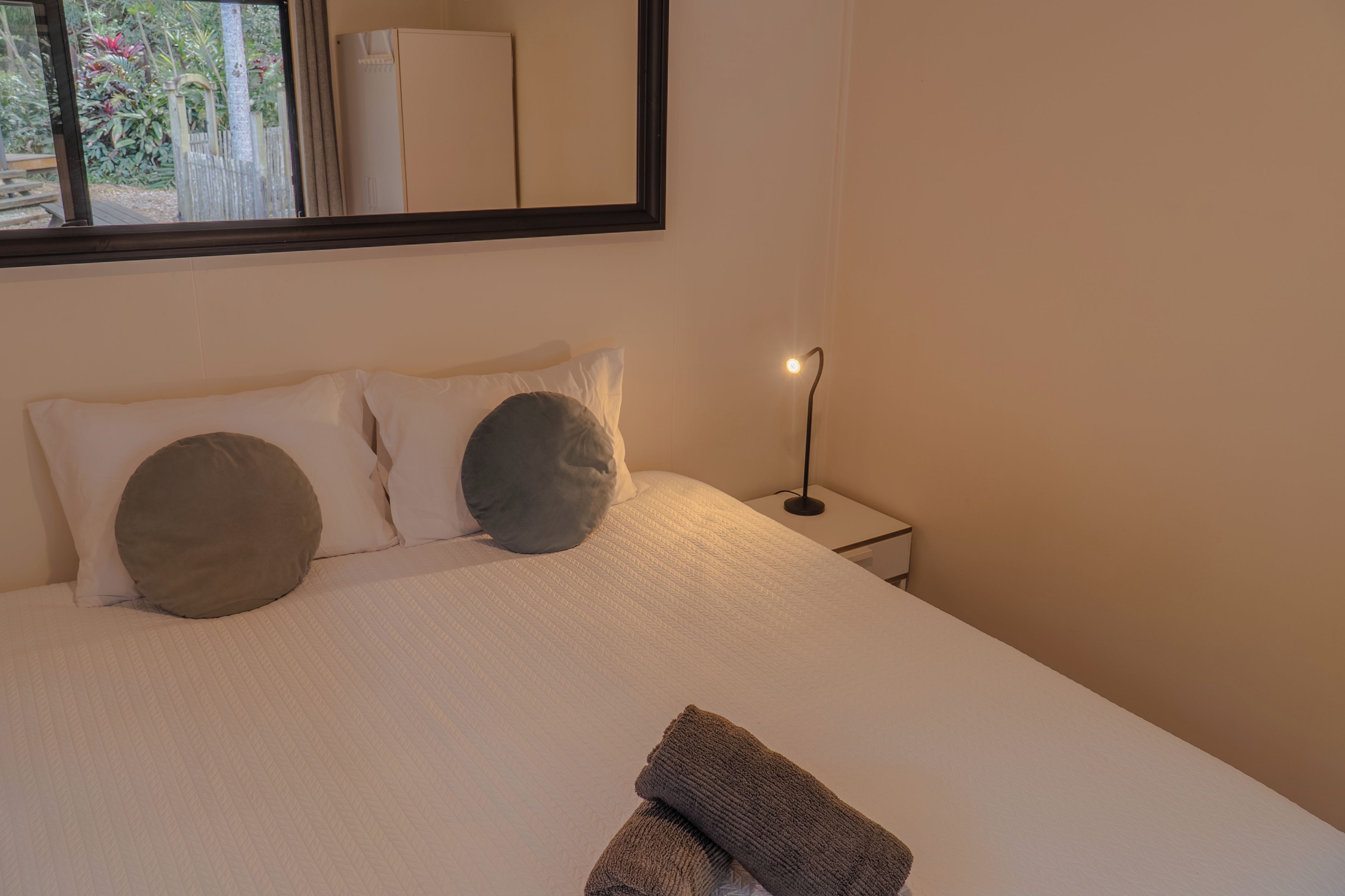 A neatly made bed with pillows and a blanket, a bedside table with a lamp, and a large mirror on the wall.
