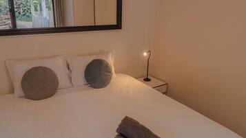 A neatly made bed with pillows and a blanket, a bedside table with a lamp, and a large mirror on the wall.