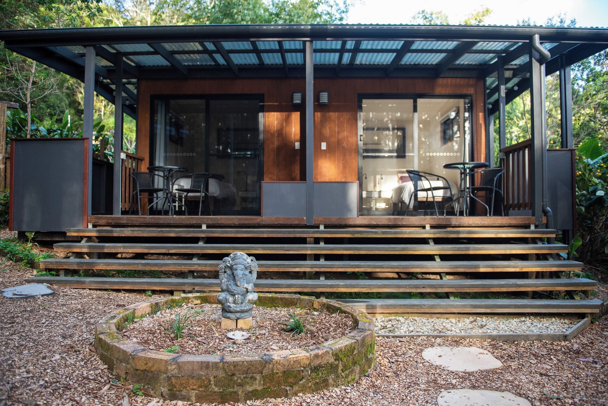 A modern building with a covered outdoor seating area, steps leading to a door, and a stone sculpture in the foreground.