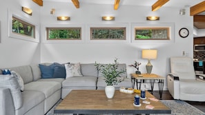 A modern living room with a sectional sofa, a coffee table, a side table with a lamp, a chair, and framed pictures on the wall. - The Grey Piper by Avantstay Ocean Views + Fire Pit + BBQ Walk to Beach (Arch Cape)