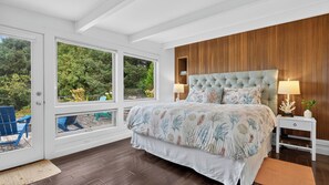 A bedroom with a large bed, two bedside lamps, a wooden headboard, and a view of the outdoors through large windows. - The Grey Piper by Avantstay Ocean Views + Fire Pit + BBQ Walk to Beach (Arch Cape)