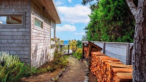 A wooden building with a clear view of the ocean, a gravel path, and stacks of wood. - The Grey Piper by Avantstay Ocean Views + Fire Pit + BBQ Walk to Beach (Arch Cape)