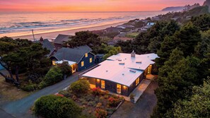 Une maison moderne avec un toit plat, entourée d’une végétation luxuriante et offrant une vue sur l’océan au coucher du soleil.